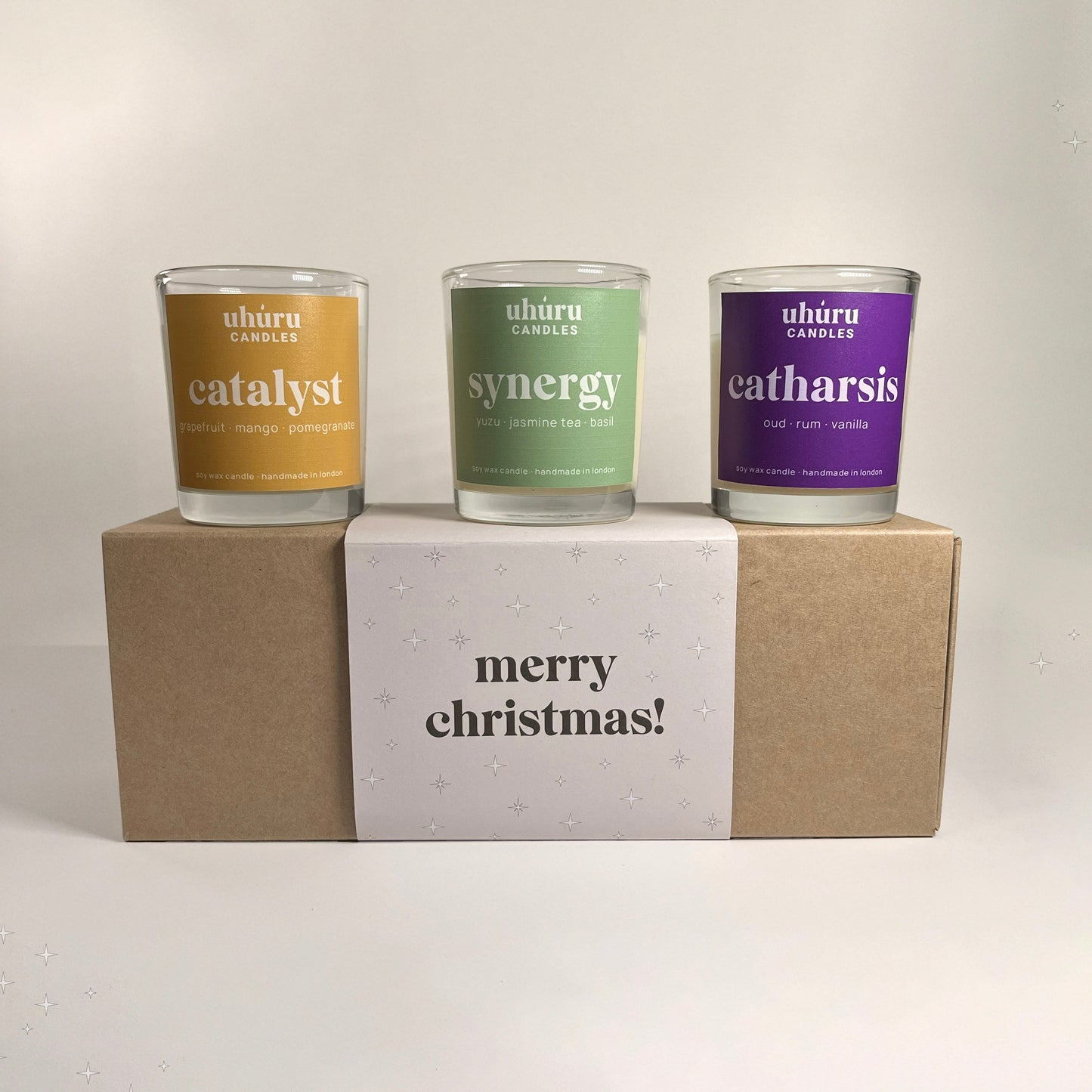 Uhuru Candles aromatherapy gift set. Three candles in glass jars labeled 'catalyst', 'synergy', and 'catharsis' on a cardboard box with 'merry christmas!' text.